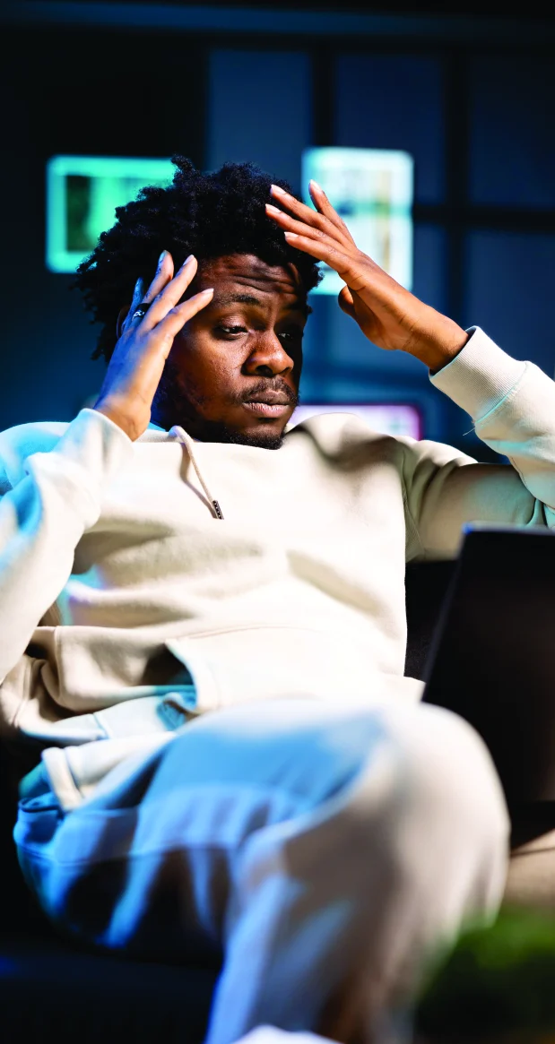 A young man holding his head, overwhelmed in front of glowing screens
